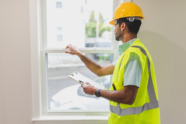 a home inspector in a yellow hard hat and vest looking at a window’s locking system